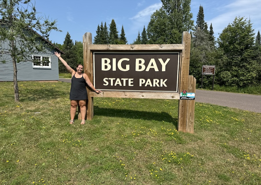 Image of young woman posing in front of entrance sign to Big Bay State Park surrounding by green grass. 